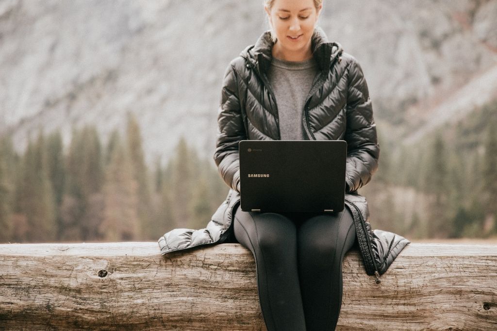 woman sitting in a wooded area with a mountain behind her working on a Samsung Chromebook